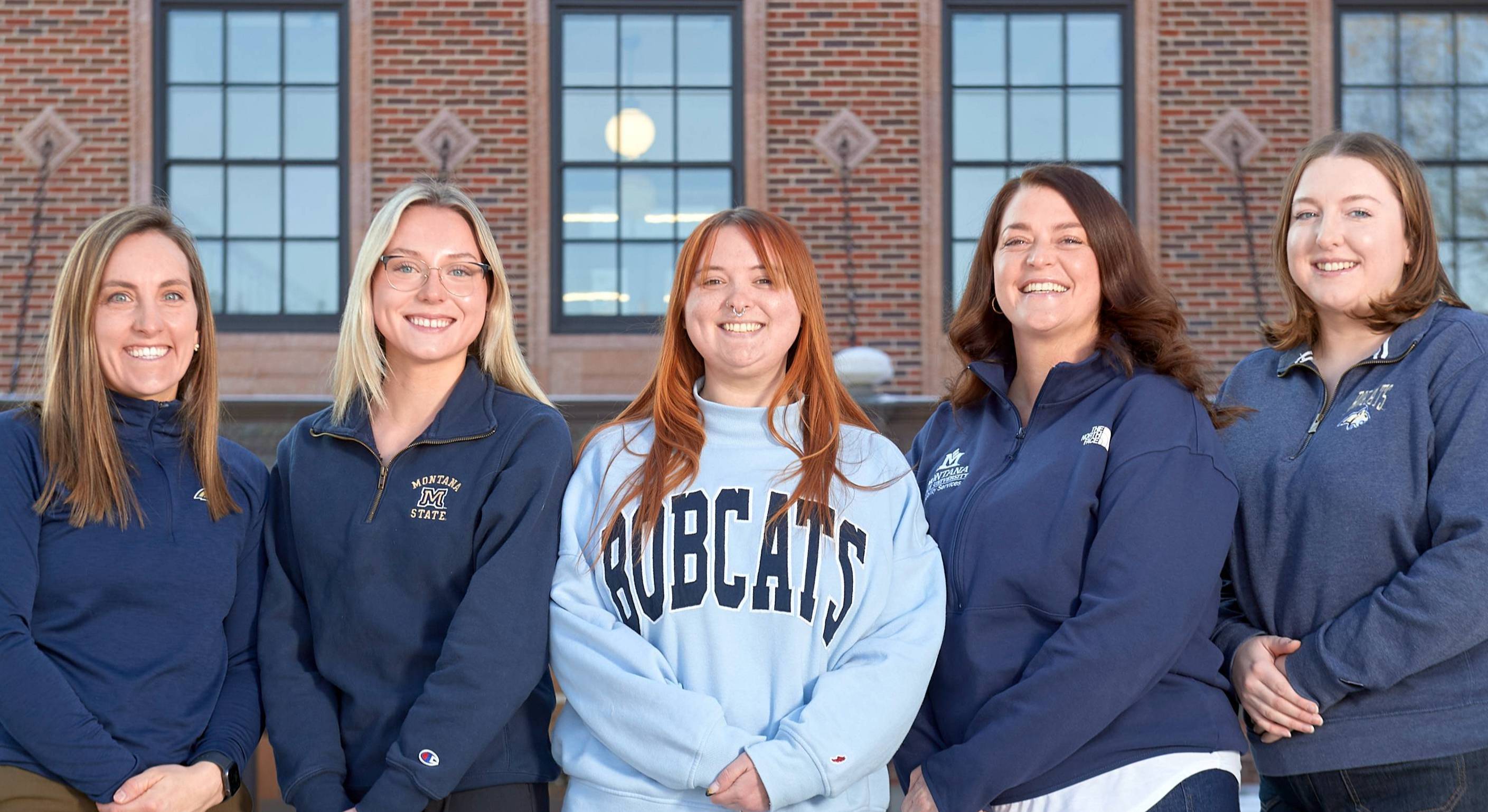 Photo of five women wearing blue Montana State University apparel. In the background is a brown brick building.