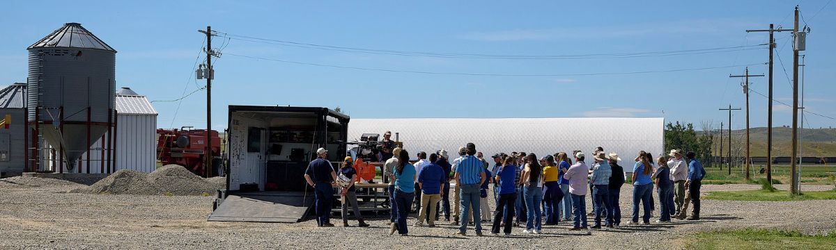 Group of people gathered outdoors around a person speaking near farm equipment and grain storage structures.