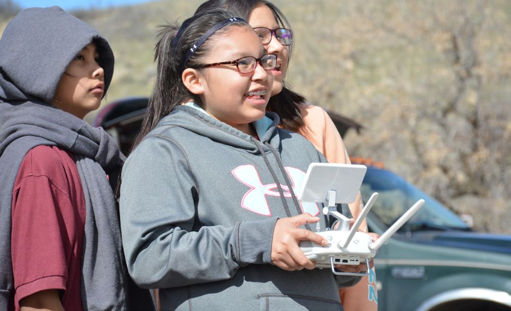 Student operating a drone controller during an outdoor educational field activity with other students nearby.