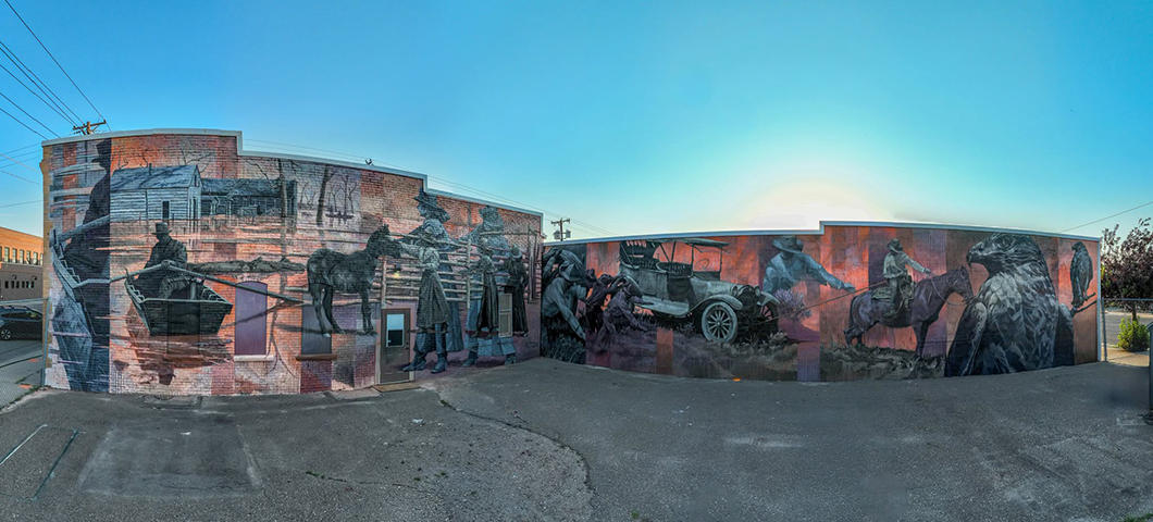 Panoramic mural painted on the exterior walls of a brick building. The artwork depicts scenes from local history, including a boat, horse riders, workers, an early automobile, and a large bird, rendered in muted blue, gray, and reddish tones.
