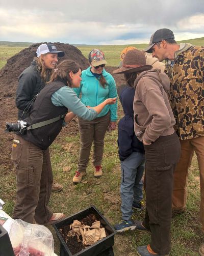 A group of people examining soil or compost material in a field demonstration while standing in open grassland.