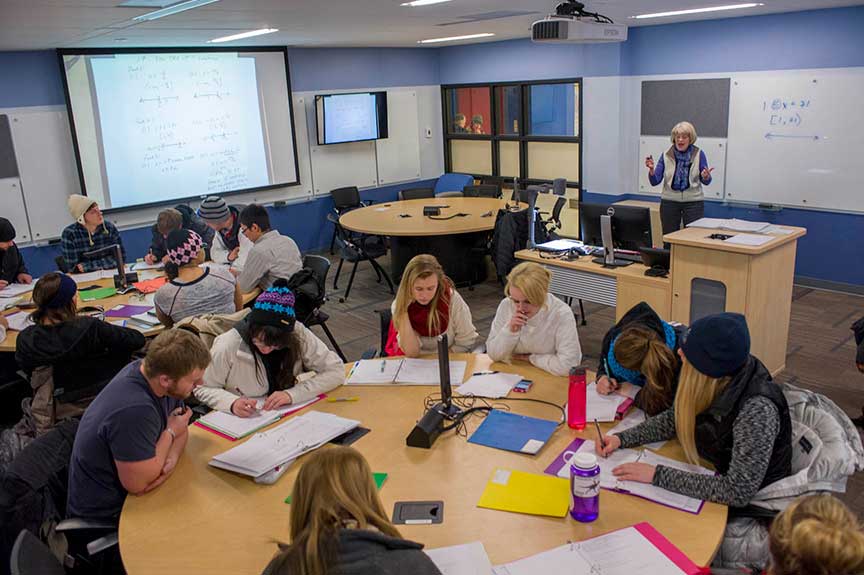 Students working around tables in a classroom with content projected on a screen and an instructor in front