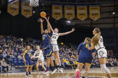 Women's college basketball player in navy blue jumps to shoot while a player in white defends under a basketball hoop.