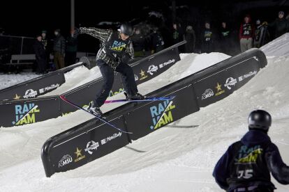 Skier performs a rail slide on a snow-covered rail during a nighttime Rail Jam event.