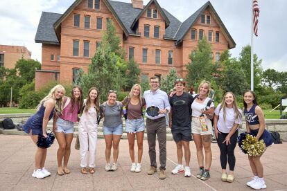 Group of students and a faculty member posing together outdoors in front of a large brick academic building.