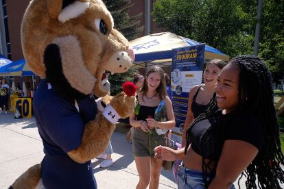 University mascot interacts and laughs with three students at an outdoor campus event with informational tents.