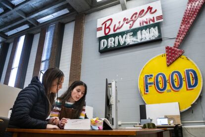 Two people study documents at a table under vintage Burger Inn Drive Inn and Food signs in an industrial-style room.