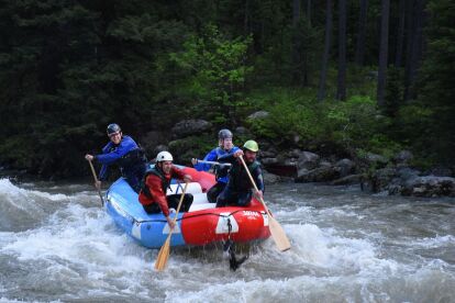 Four people wearing helmets and life jackets paddle a raft through whitewater rapids surrounded by forest.