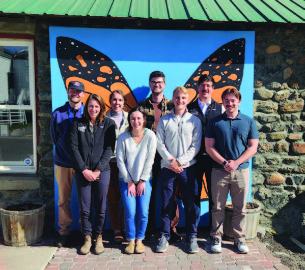 A grou of eight women and mean  standing in front of a large butterfly mural, smiling and looking straight ahead. 