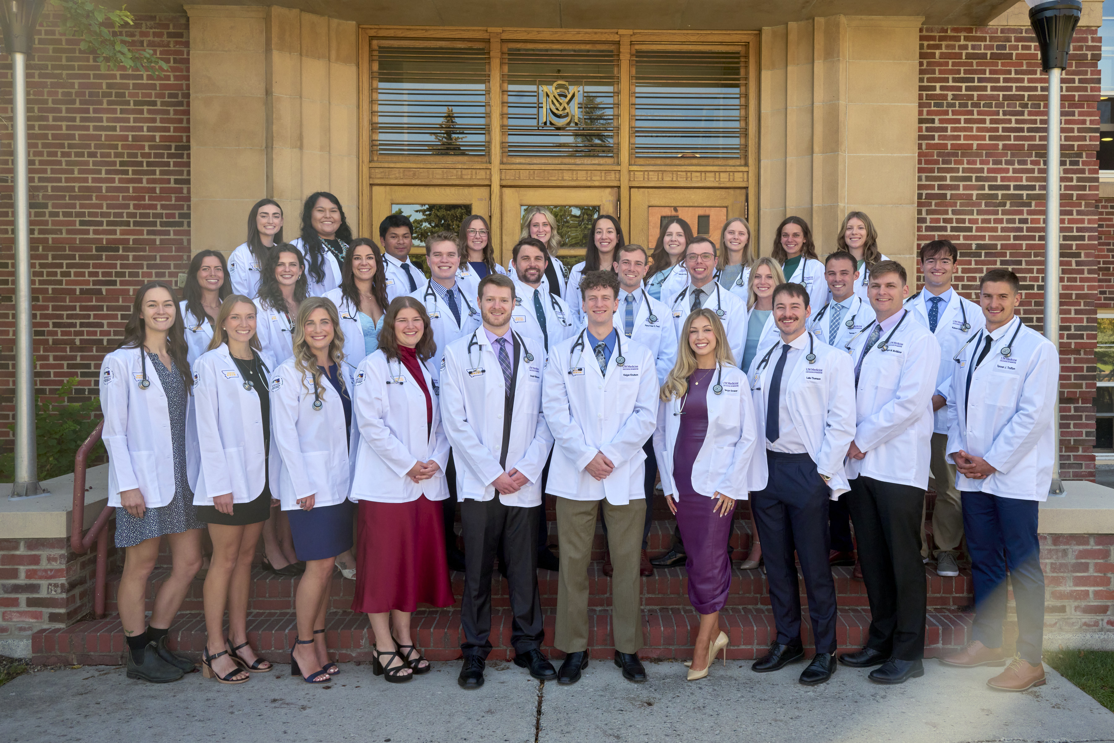 A group of 30  male and female medical students posing in their white coats in front of a brick building
