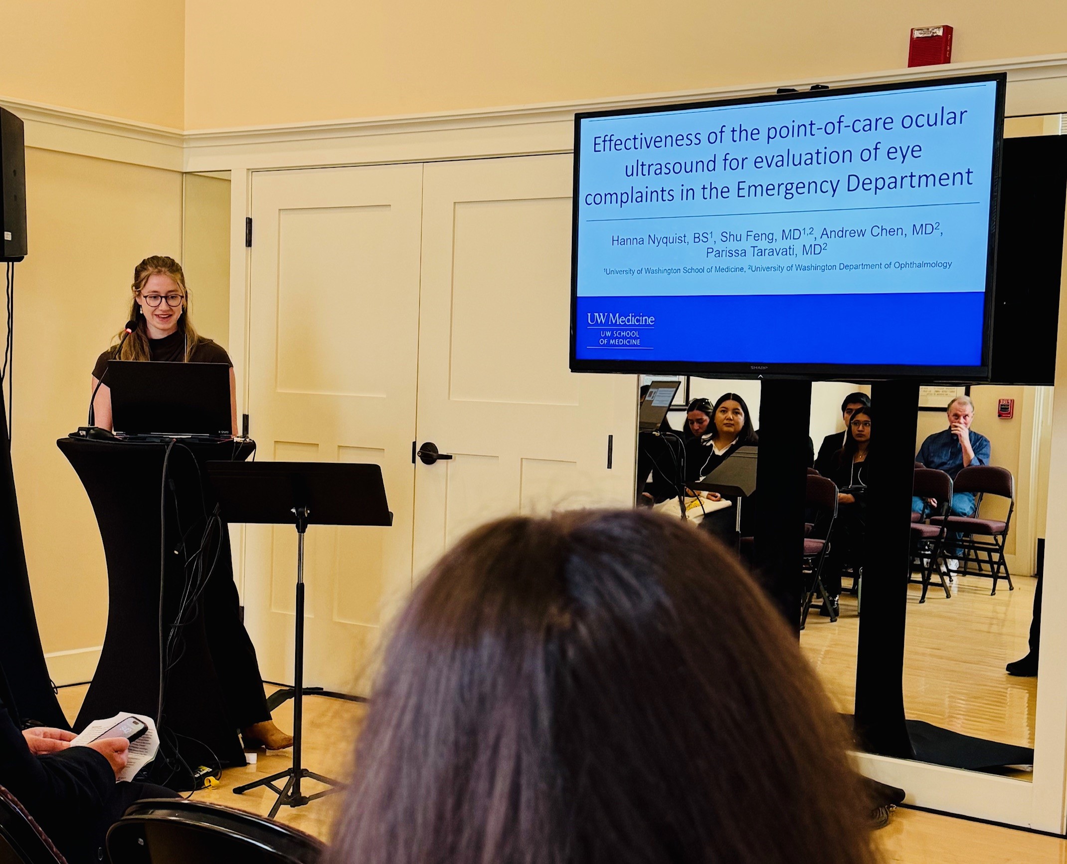 A female medical student at a podium next to a large screen showing a slide from a research presentation